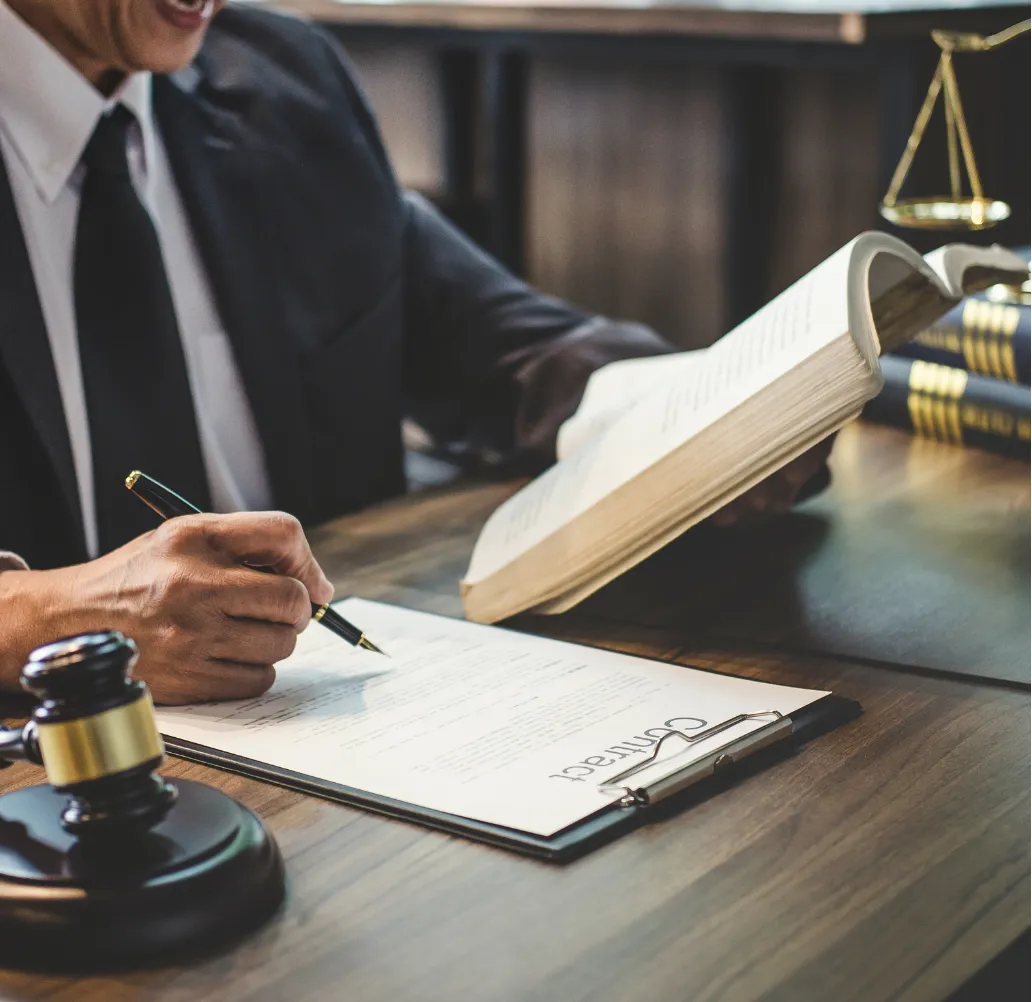 A person in a suit reviews a contract at a desk with legal books, a gavel, and scales of justice.