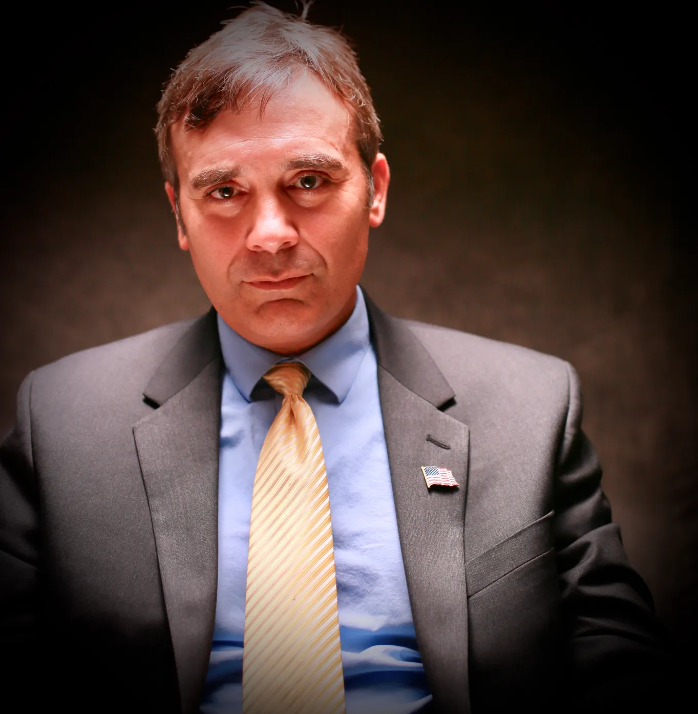 areas-2 A man in a suit with an American flag pin sits under dramatic lighting, looking directly at the camera.
