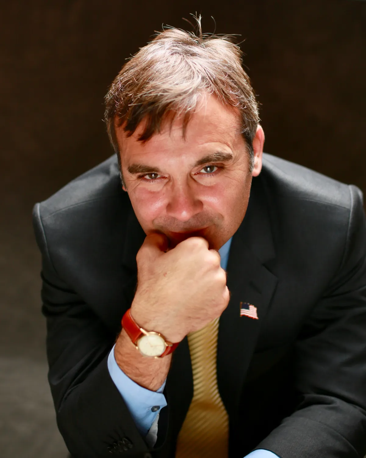 Man in suit with American flag pin, hand on chin, looking up thoughtfully against dark background.