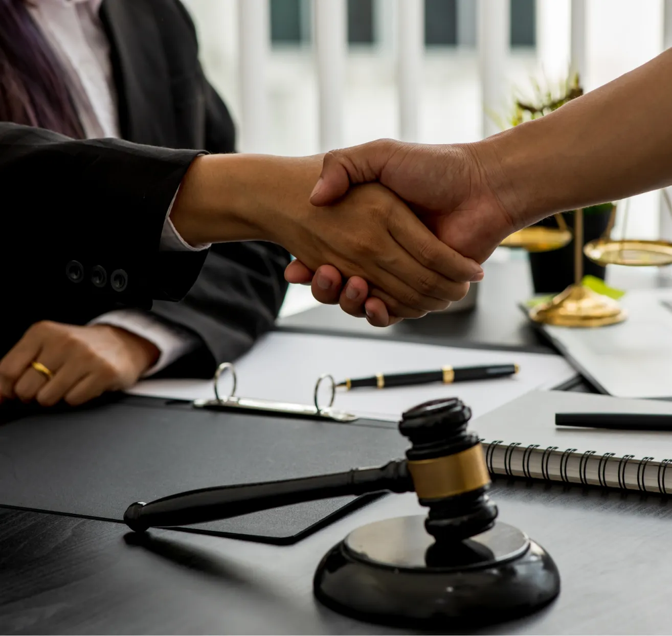 Two people in business attire shaking hands at a desk with legal documents, a gavel, and scales of justice.