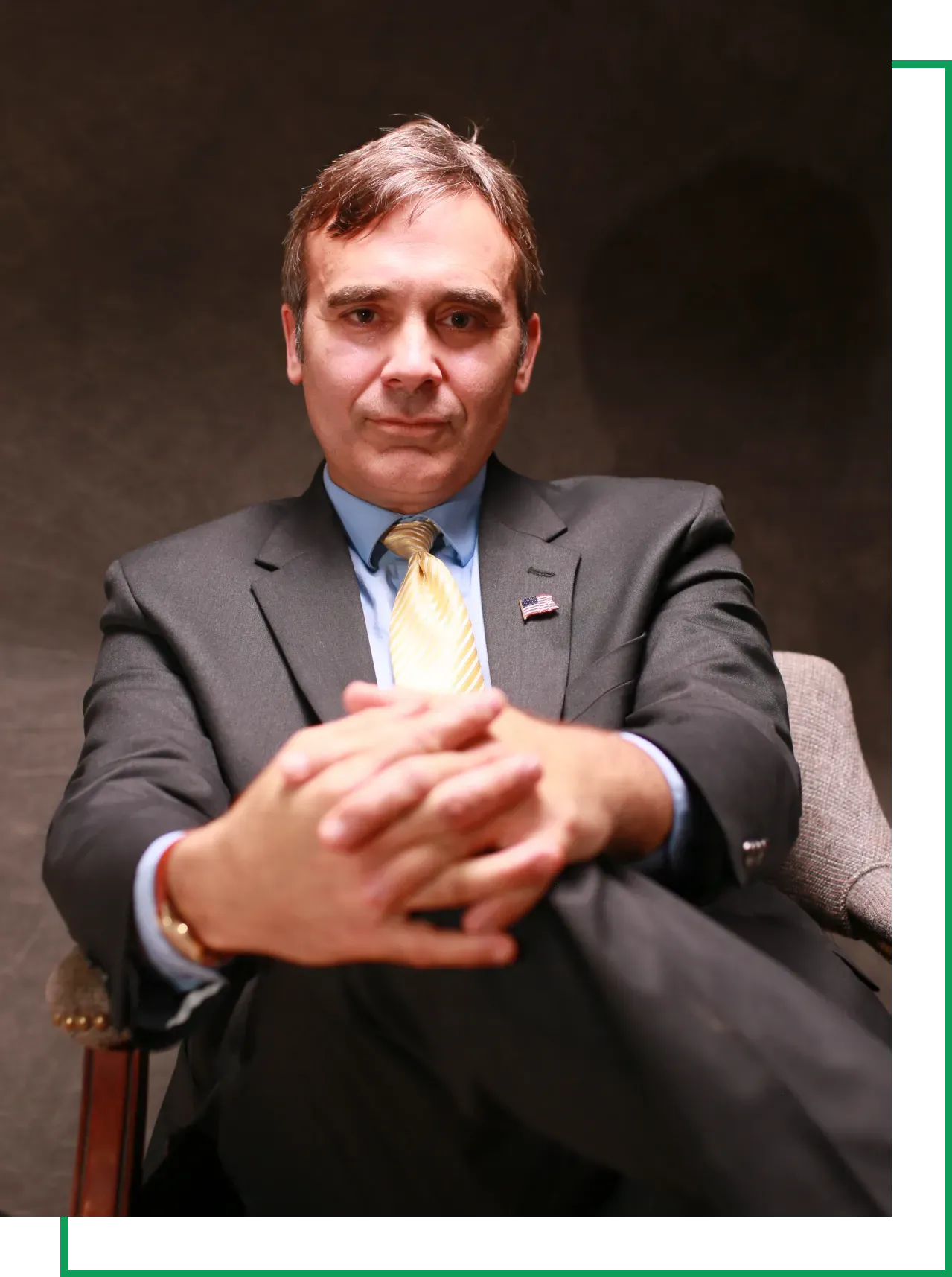 Man in a suit and yellow tie sits in a chair with hands clasped, looking confidently at the camera.